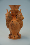 An orange glazed ceramic vase featuring two detailed goat heads with horns on either side of the neck, with floral relief patterns on the front, set against a light gray background.