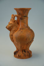 An orange-brown terracotta-style vase with a sculpted goat head handle and a flared rim, shown against a light gray studio background.