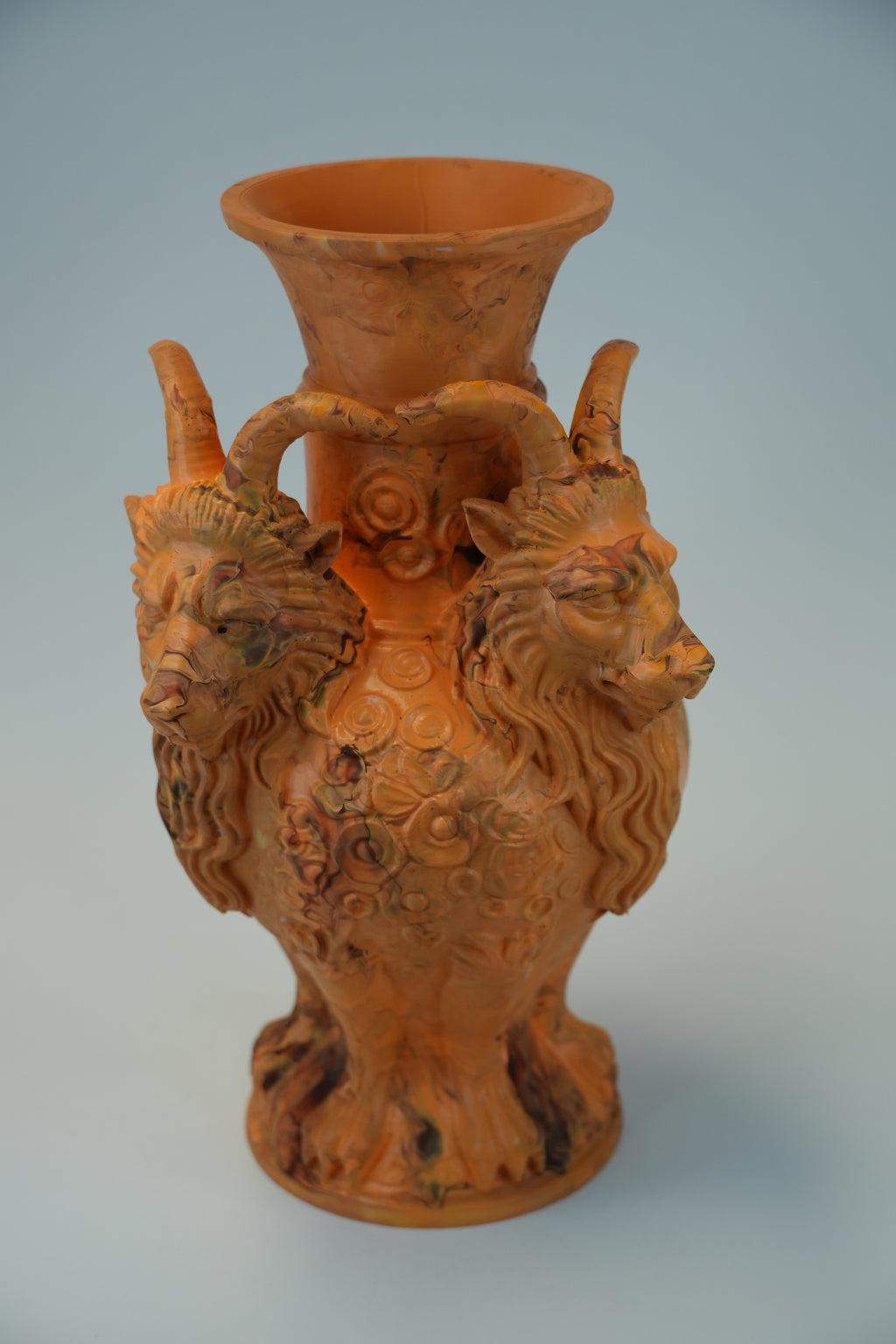 An antique terracotta ram vase with a wide flared rim, featuring two detailed ram heads on the sides and a central floral pattern, set against a light gray background.