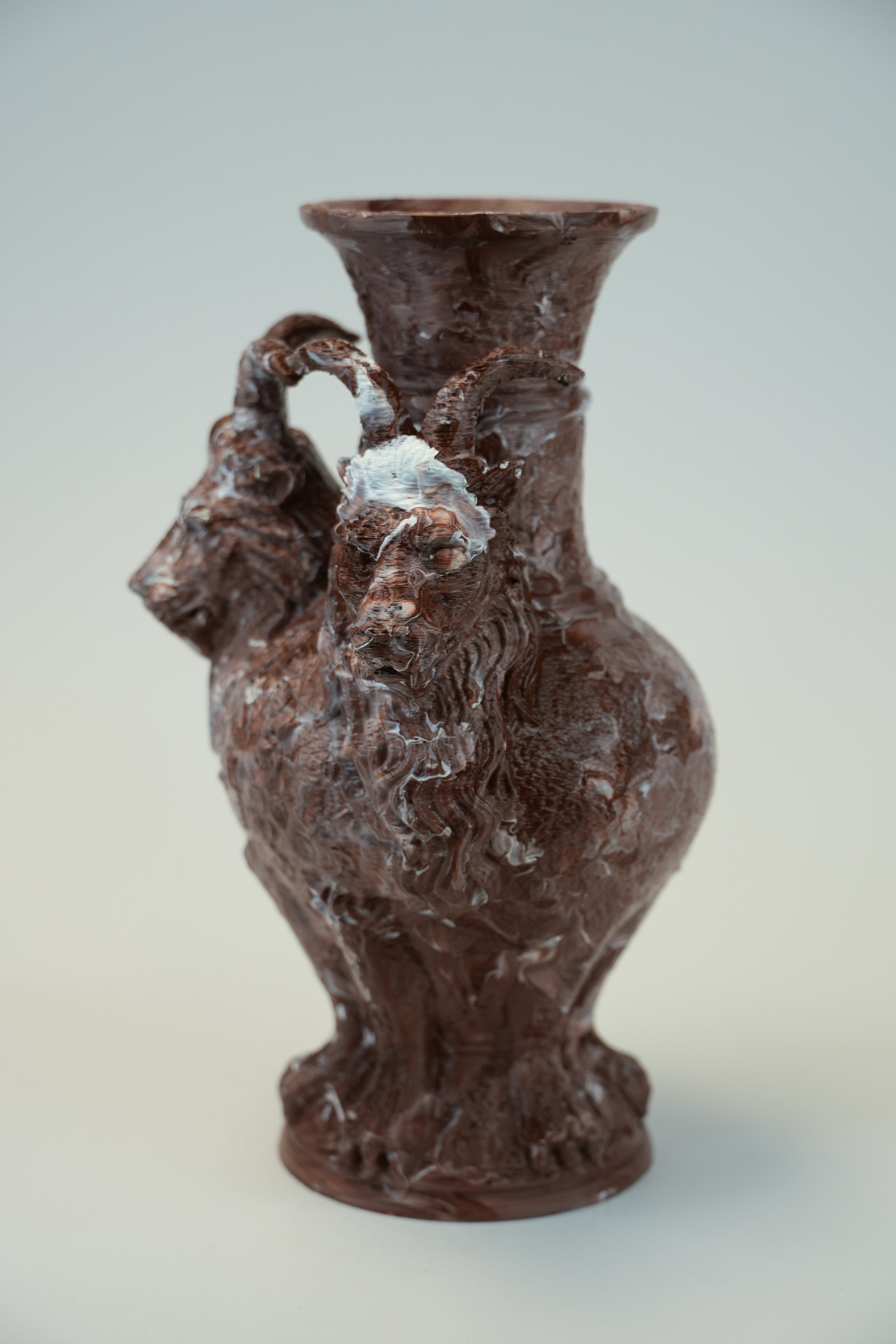 A dark brown ceramic vase with a sculpted ram head handle and a textured, marbled finish, photographed against a neutral light gray background.