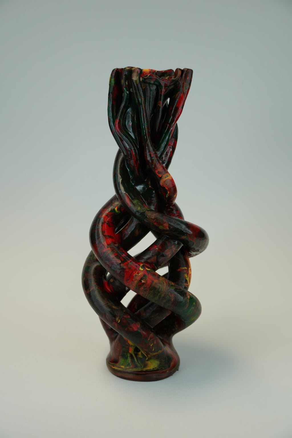 An abstract sculptural vase with a twisted, interlaced organic form, featuring a glossy dark finish with red, green, and yellow marbled accents, set against a neutral light gray background.