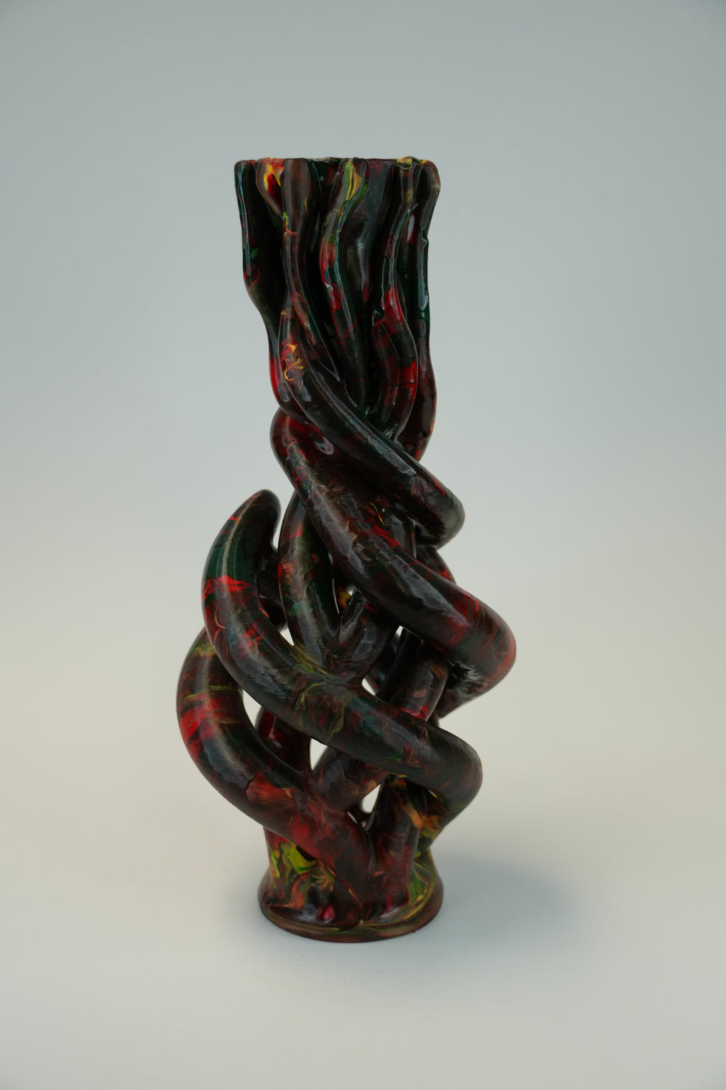 A sculptural twisted vase with a glossy finish, featuring a marbled pattern of black, deep red, and hints of green, set against a neutral light gray background.