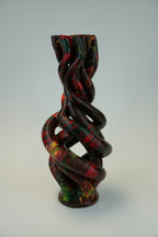 A sculptural twisted vase with a glossy finish, featuring a marbled pattern of black, red, green, and yellow, set against a plain light gray background.
