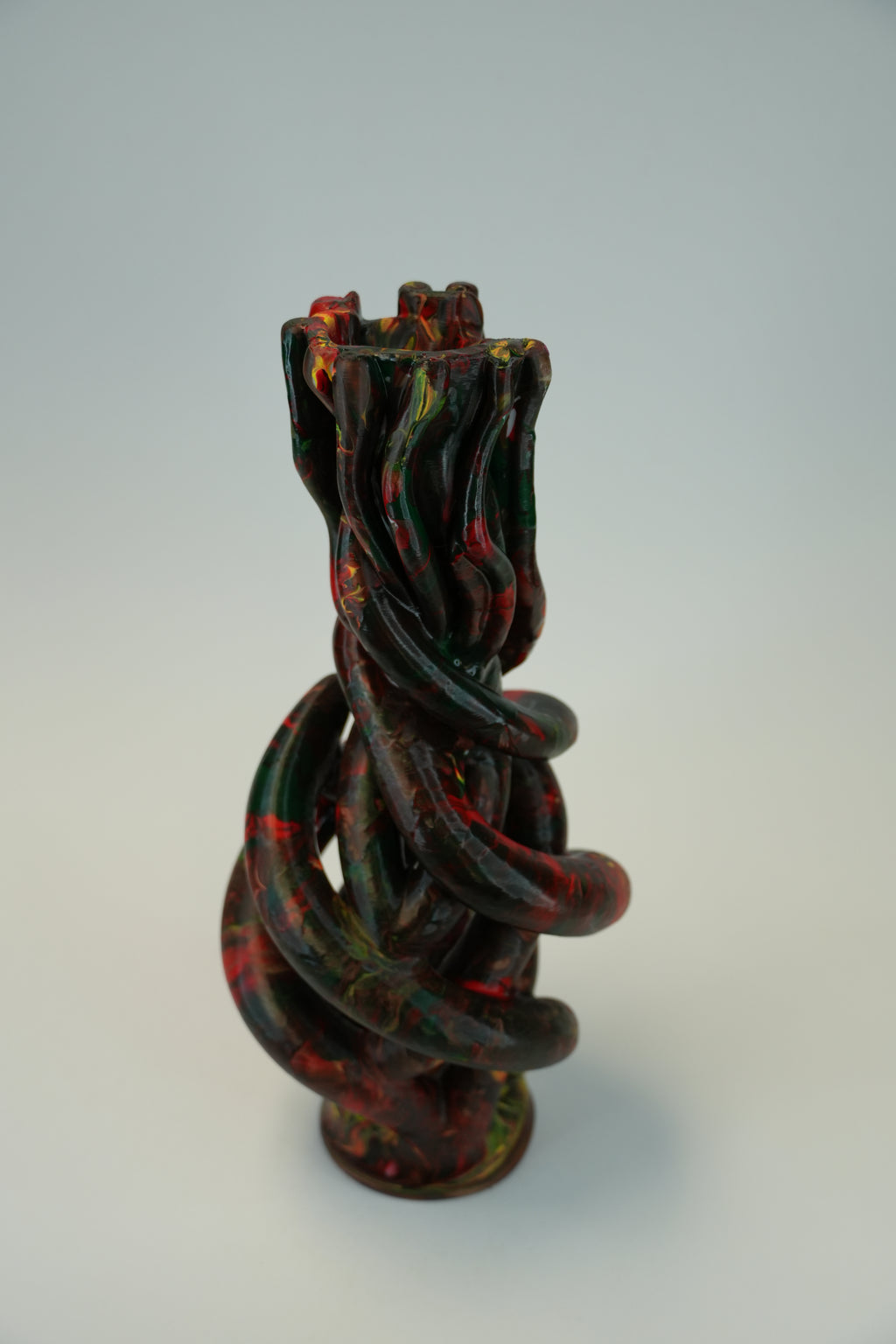 A sculptural twisted vase with a glossy finish, featuring a marbled pattern of black, deep green, and red, set against a plain light gray background.