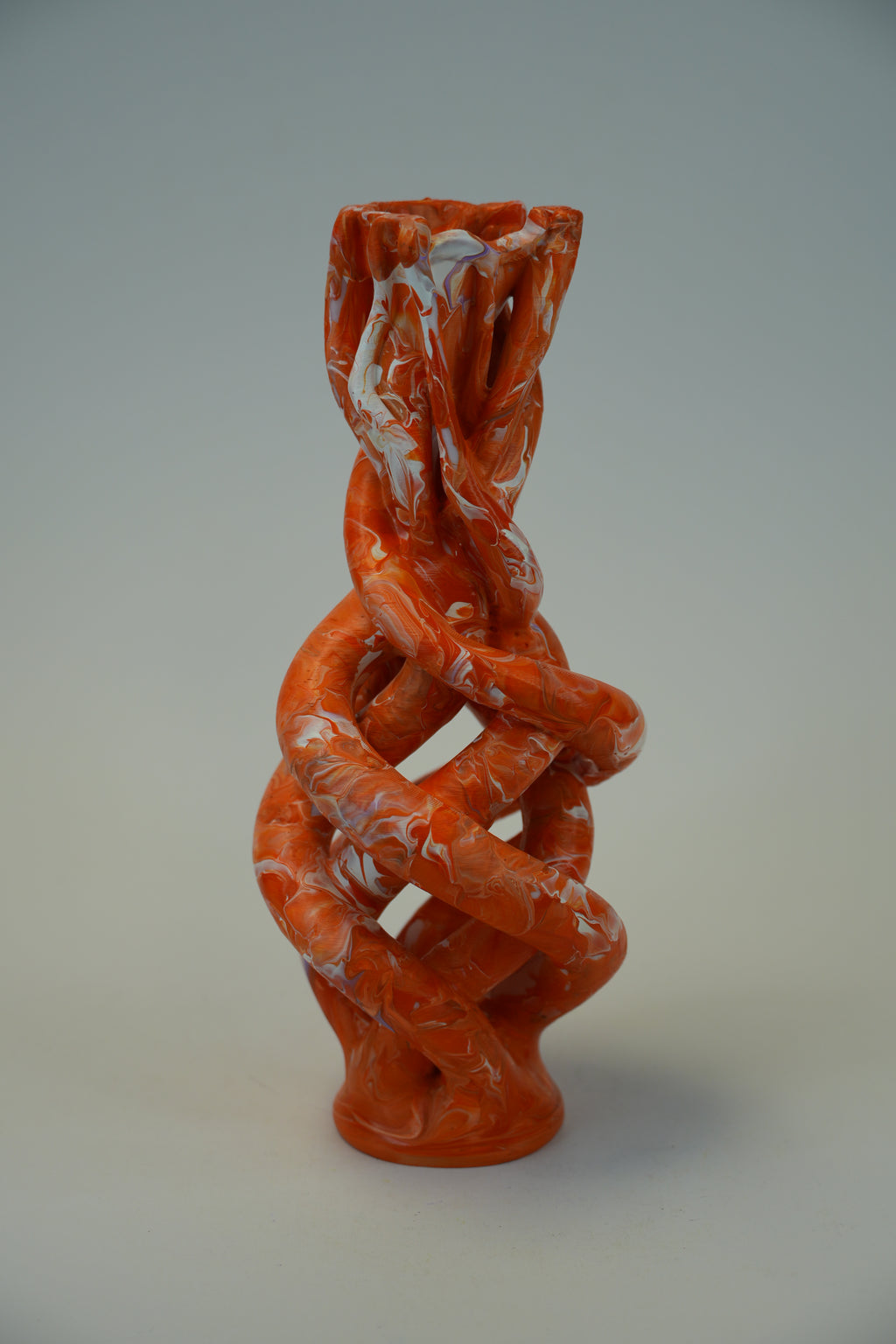An abstract sculptural vase with a twisted, interlaced organic form in a bright orange-red glaze with white marbled streaks, photographed against a neutral light gray studio background.