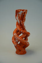 An abstract sculptural vase with a twisted, interlaced organic form in a bright orange-red glaze with white marbled streaks, photographed against a neutral light gray studio background.