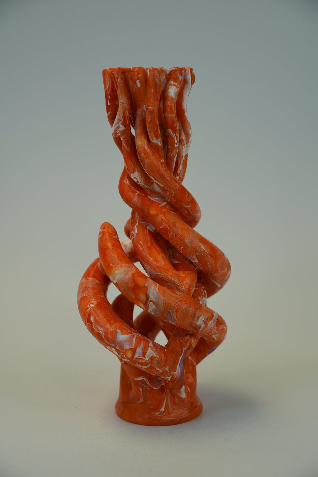 An abstract sculptural vase with a twisted, organic spiral form in glossy terracotta orange with white marbled streaks, set against a neutral light gray background.