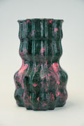 A sculptural, ribbed ceramic vase with a glossy finish, featuring a marbled pattern of dark green and vibrant pink, set against a plain light gray background.