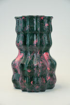 A sculptural, ribbed ceramic vase with a glossy finish, featuring a marbled pattern of dark green and vibrant pink, set against a plain light gray background.