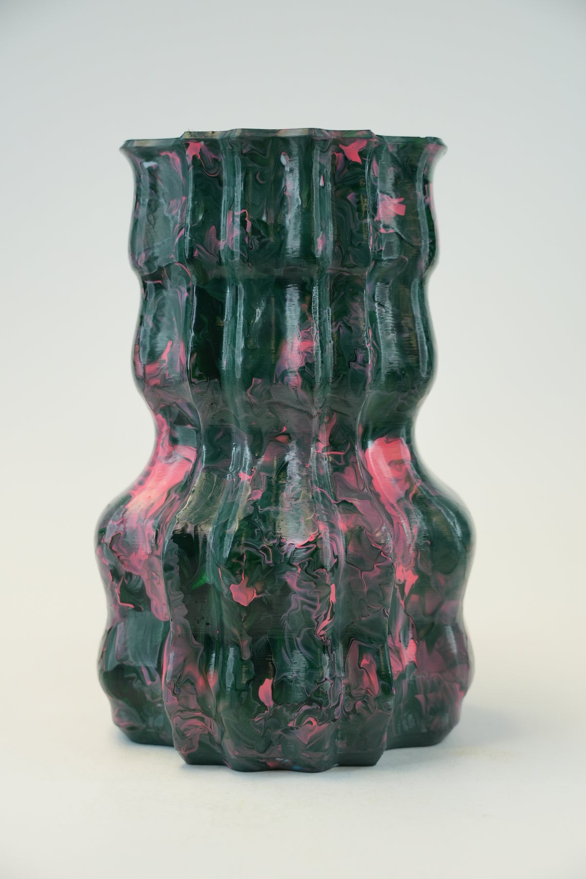A sculptural, ribbed ceramic vase with a glossy finish, featuring a marbled pattern of dark green and vibrant pink, set against a plain light gray background.