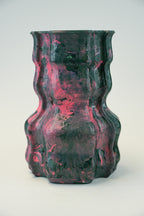 A sculptural, wavy ceramic vase with a marbled pattern of dark green and vibrant pink, photographed against a neutral light gray background.