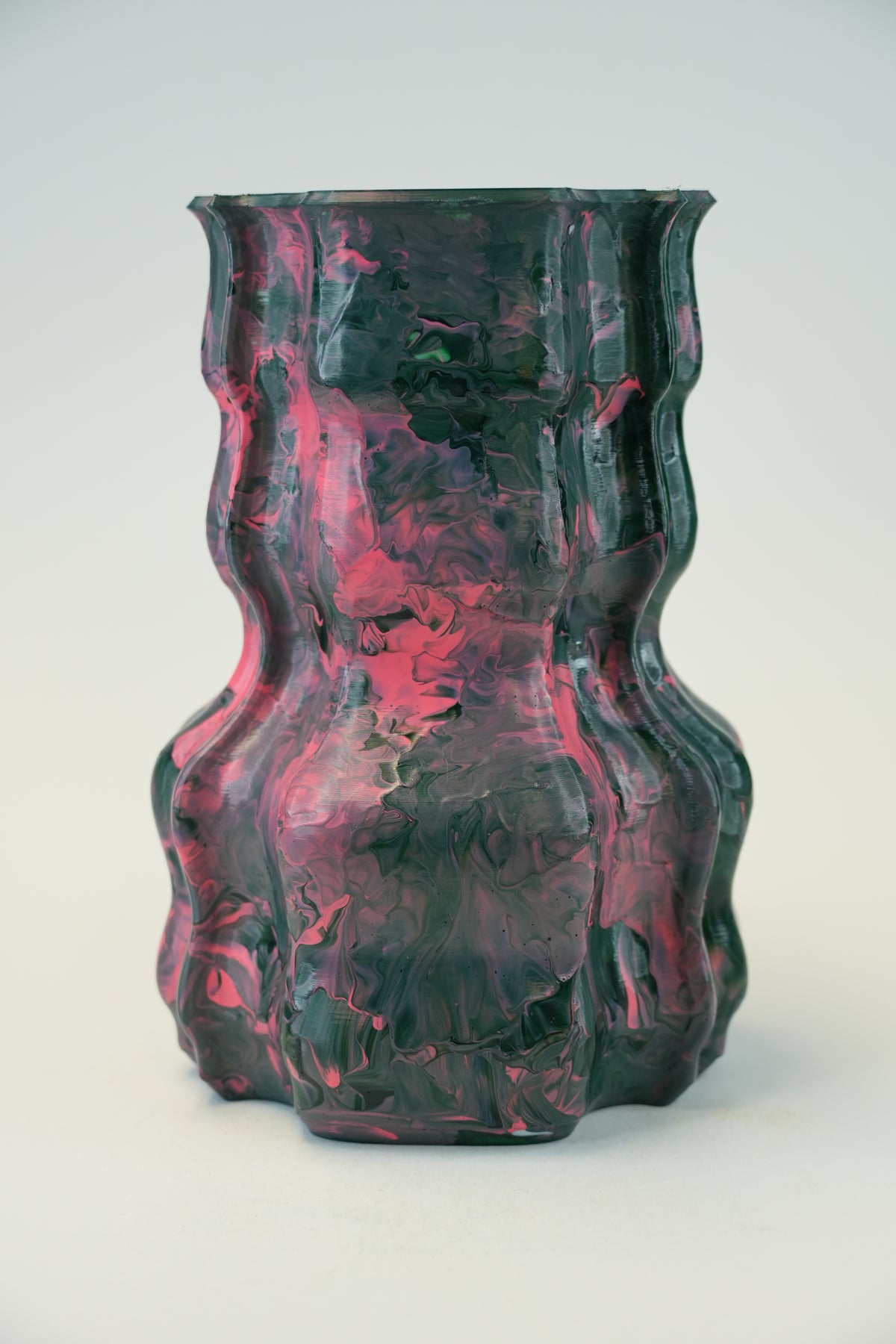 A sculptural, wavy ceramic vase with a marbled pattern of dark green and vibrant pink, photographed against a neutral light gray background.