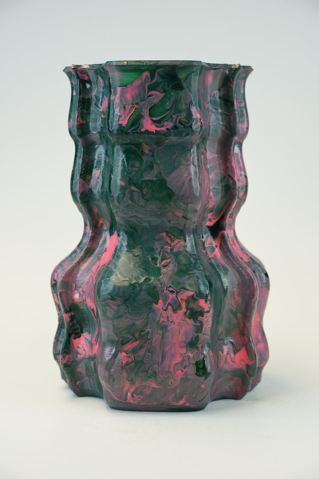 A sculptural, wavy glass vase with a marbled pattern of dark green, black, and vibrant pink, photographed on a seamless light gray background.