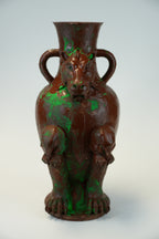 A sculptural ceramic vase shaped like a seated tiger, with two side handles and a glossy finish, featuring a dark brown base color with vibrant green paint splatter accents, set against a plain light gray background.