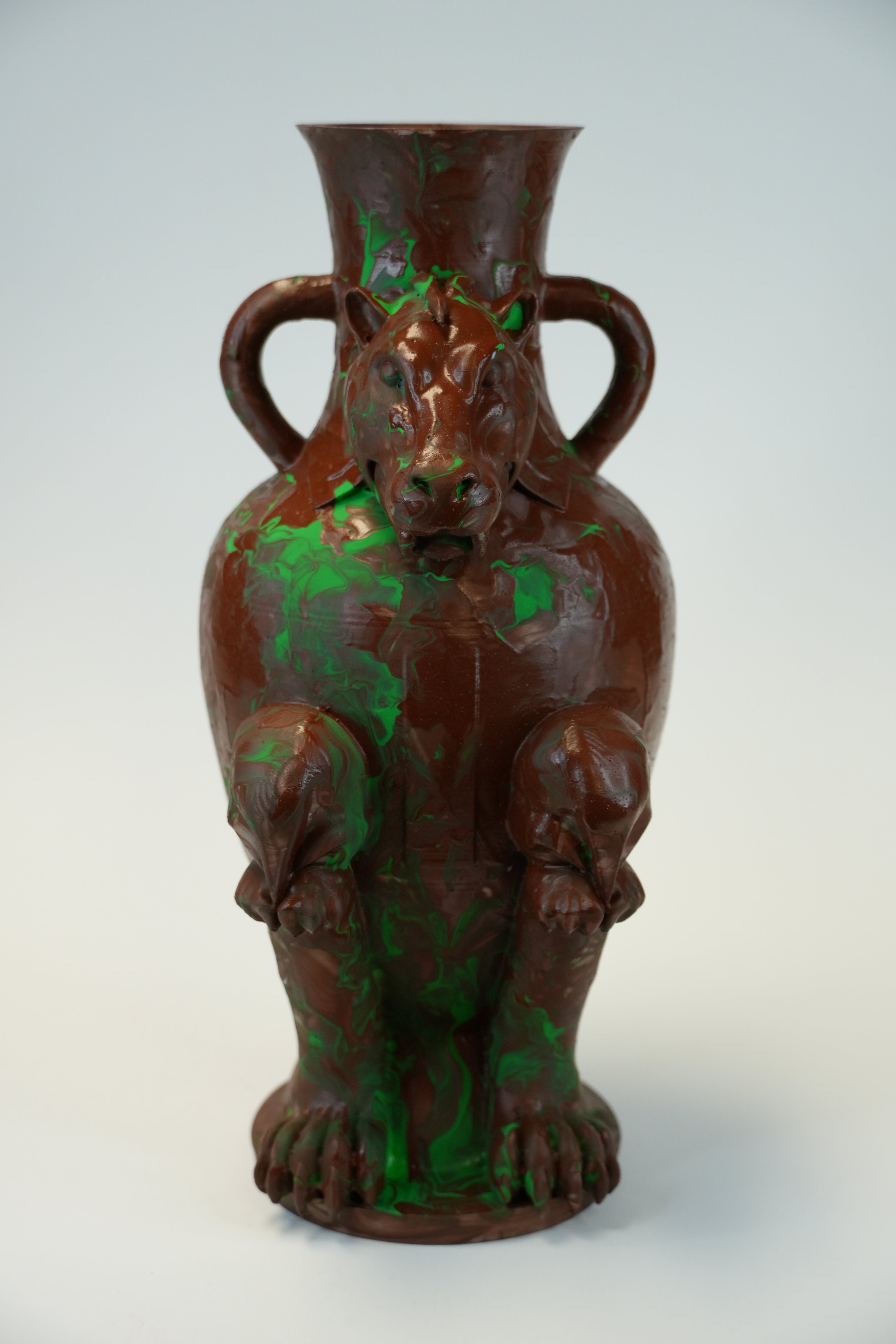 A sculptural ceramic vase shaped like a seated tiger, with two side handles and a glossy finish, featuring a dark brown base color with vibrant green paint splatter accents, set against a plain light gray background.