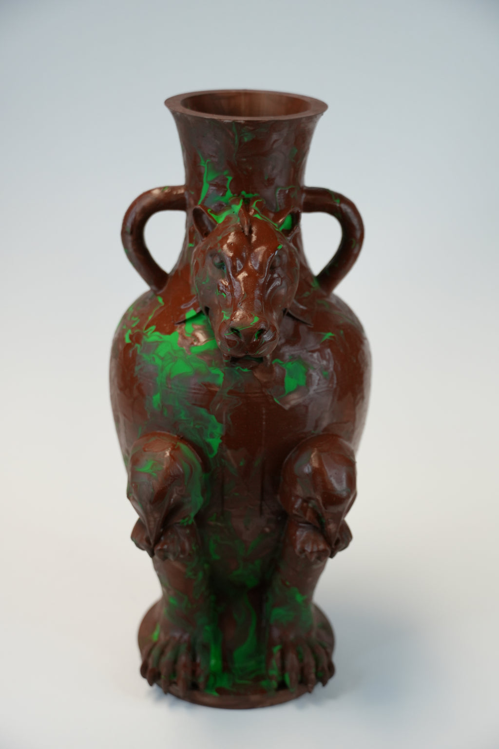 A sculptural ceramic vase shaped like a seated bear, with two side handles and a flared rim, shown on a plain light gray studio background. The surface is predominantly dark brown with irregular bright green painted accents.