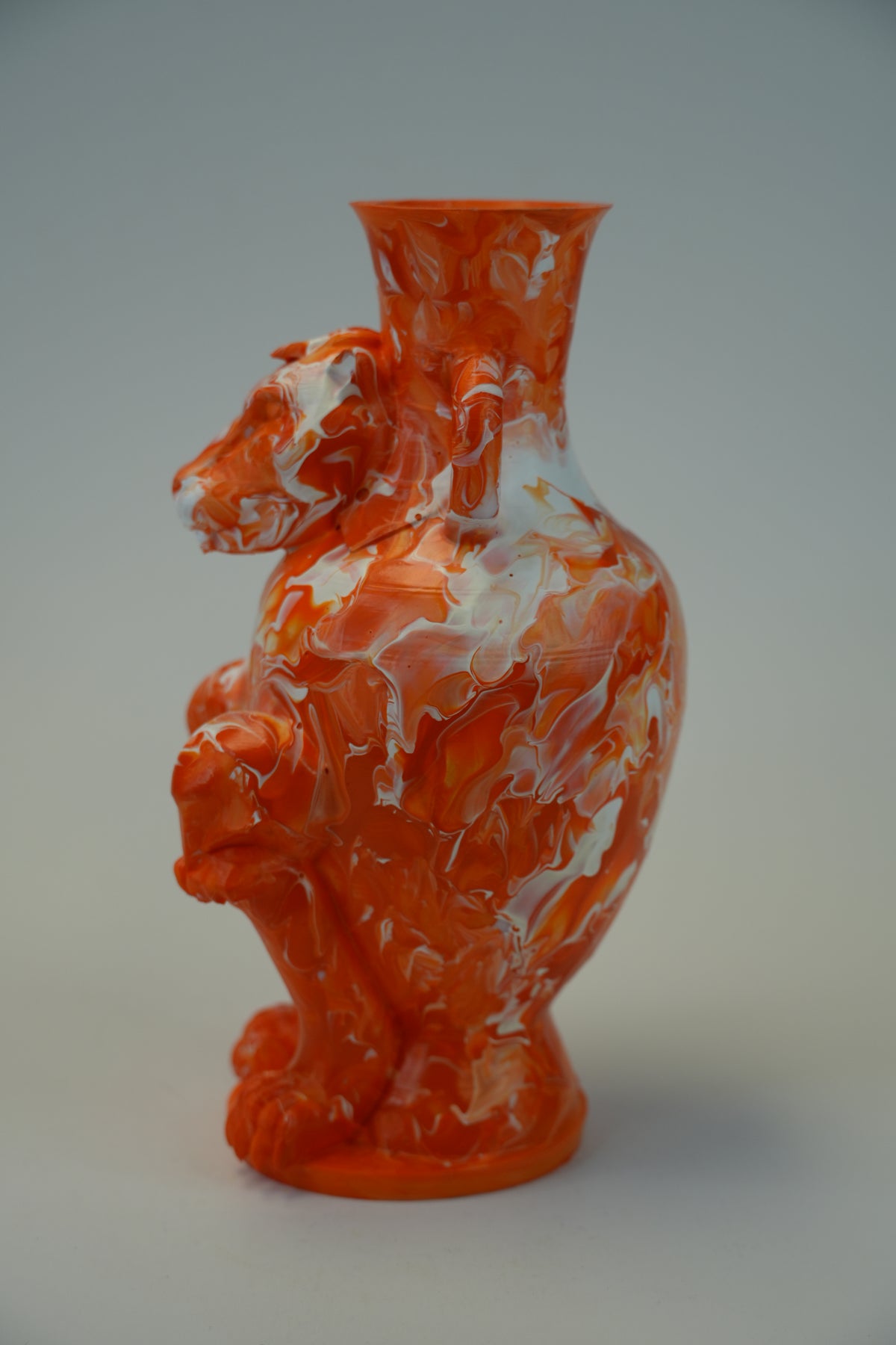 An orange sculptural vase with a hippo figure integrated into the body, featuring a marbled swirl pattern of white and orange, set against a neutral gray background.