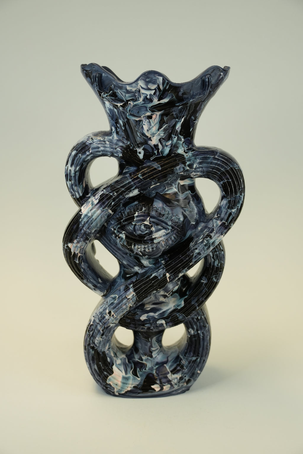 A sculptural glass vase with a dark blue and white marbled pattern, featuring an eye motif in the center and a flared rim, set against a neutral light gray background.