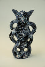 A sculptural glass vase with a dark blue and white marbled pattern, featuring an eye motif in the center and a flared rim, set against a neutral light gray background.