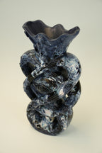 An abstract sculptural ceramic vase with a flared irregular rim, featuring a marbled swirl pattern in dark blue, black, and white, set against a neutral light gray background.