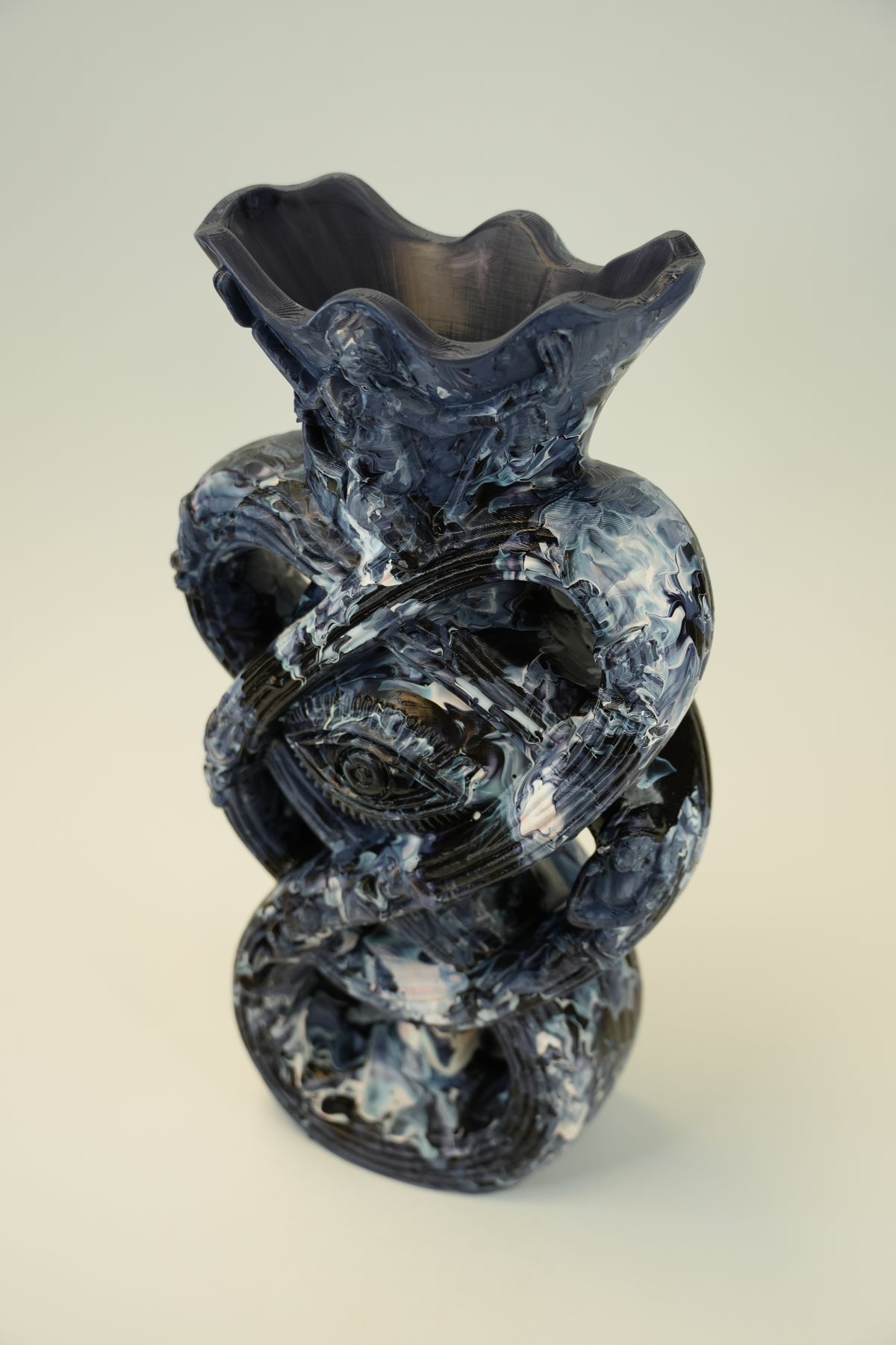 An abstract sculptural ceramic vase with a flared irregular rim, featuring a marbled swirl pattern in dark blue, black, and white, set against a neutral light gray background.