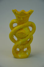 A bright yellow sculptural ceramic vase with a flared rim, ribbed texture, and an eye motif wrapped through the center, photographed against a neutral light gray studio background.