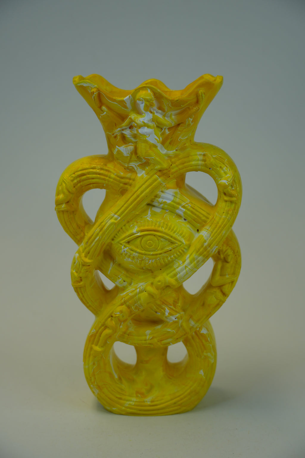 A yellow sculptural vase with an abstract, organic form featuring a central eye motif and looped, snake-like bands, photographed against a neutral gray studio background.