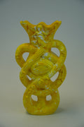 A yellow sculptural vase with an abstract, organic form featuring a central eye motif and looped, snake-like bands, photographed against a neutral gray studio background.
