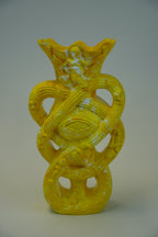 A yellow sculptural vase with an abstract, organic form featuring a central eye motif and looped, snake-like bands, photographed against a neutral gray studio background.