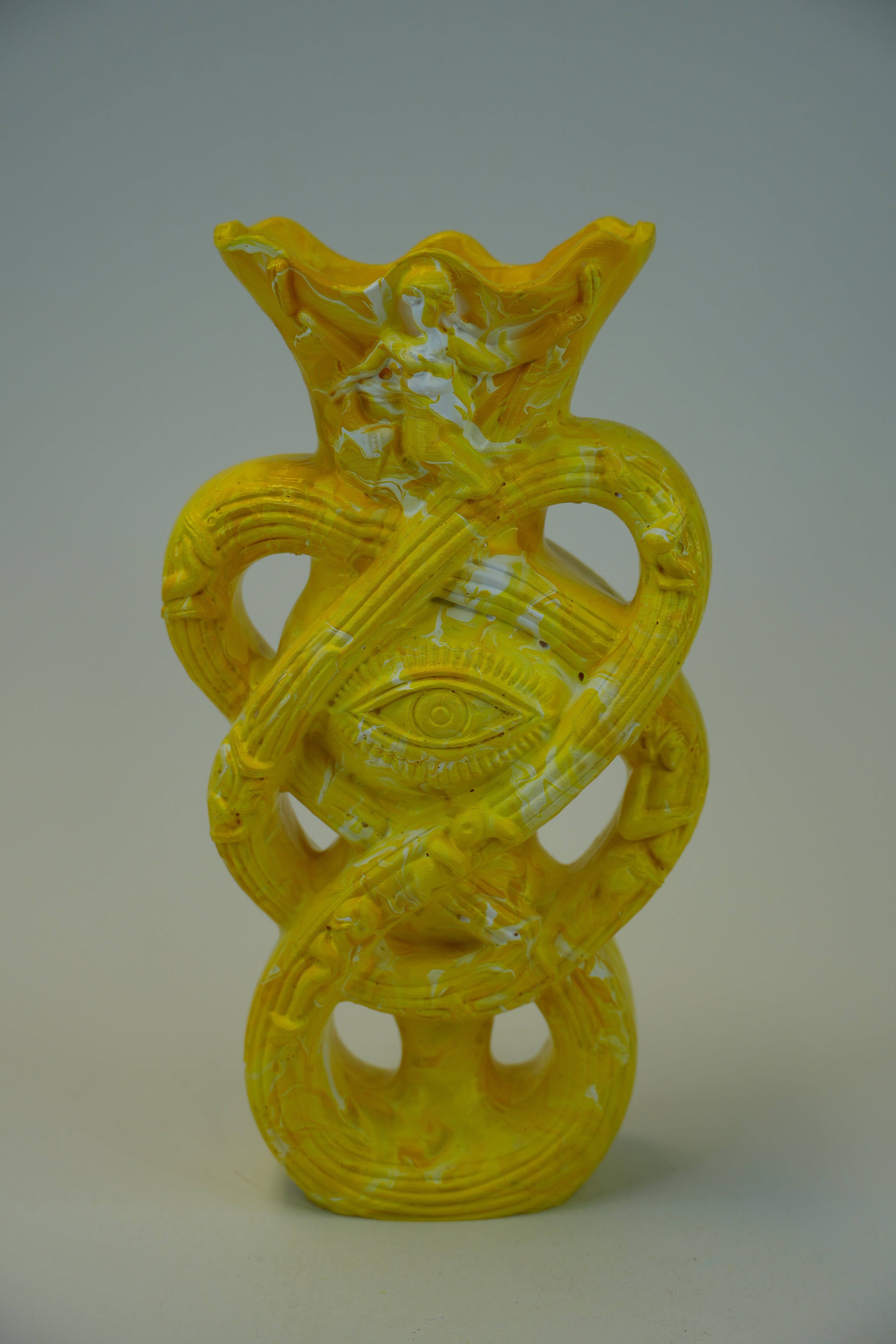 A yellow sculptural vase with an abstract, organic form featuring a central eye motif and looped, snake-like bands, photographed against a neutral gray studio background.