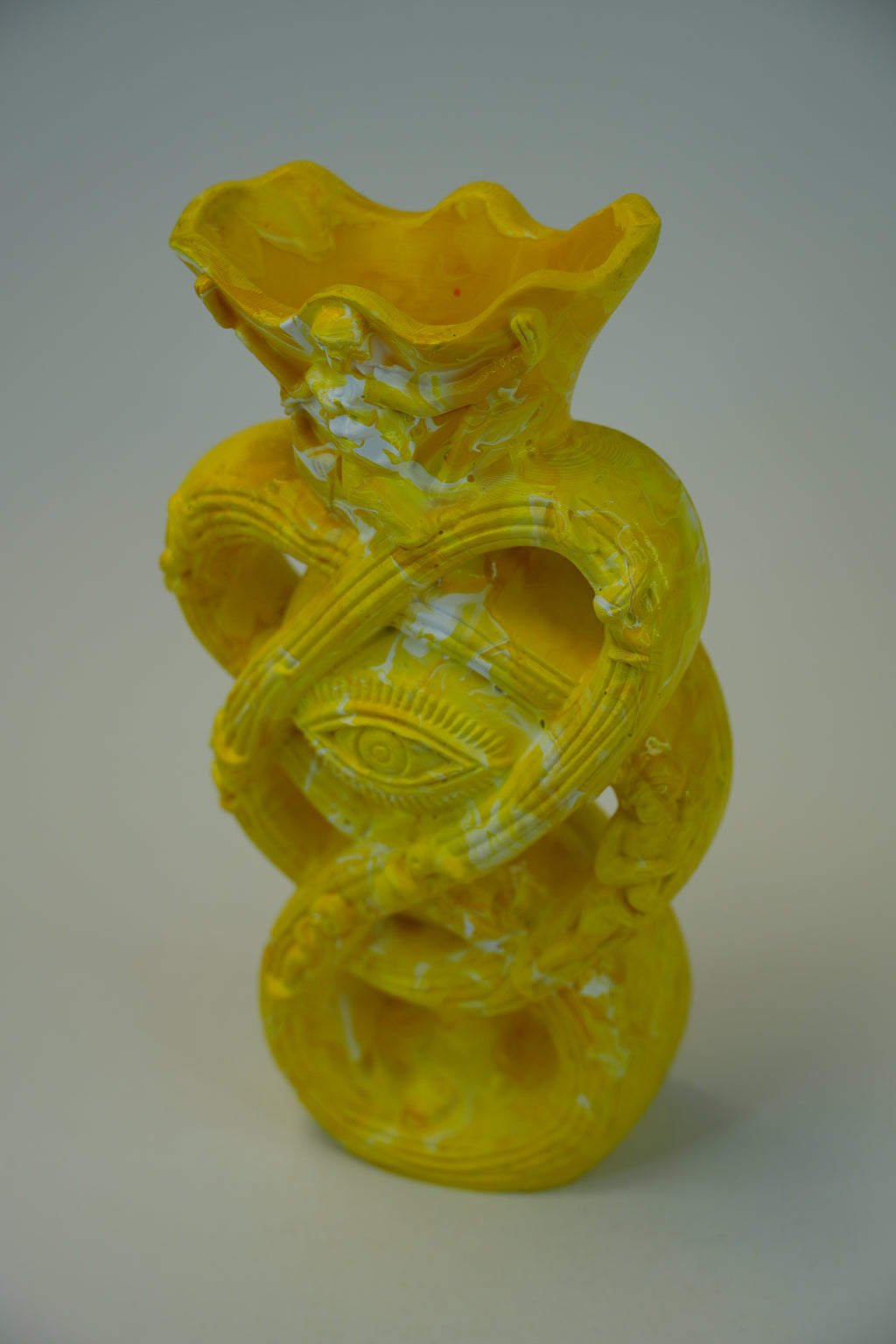 A bright yellow sculptural ceramic vase with a textured, hand-painted finish and a central eye motif, set against a neutral light gray background.