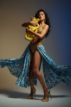 A woman poses in a gold bikini and flowing blue patterned sarong against a studio backdrop, holding a large yellow snake.