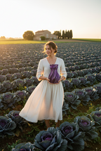 Woman holding a purple vase in a field of cabbages with a sunset in the background.