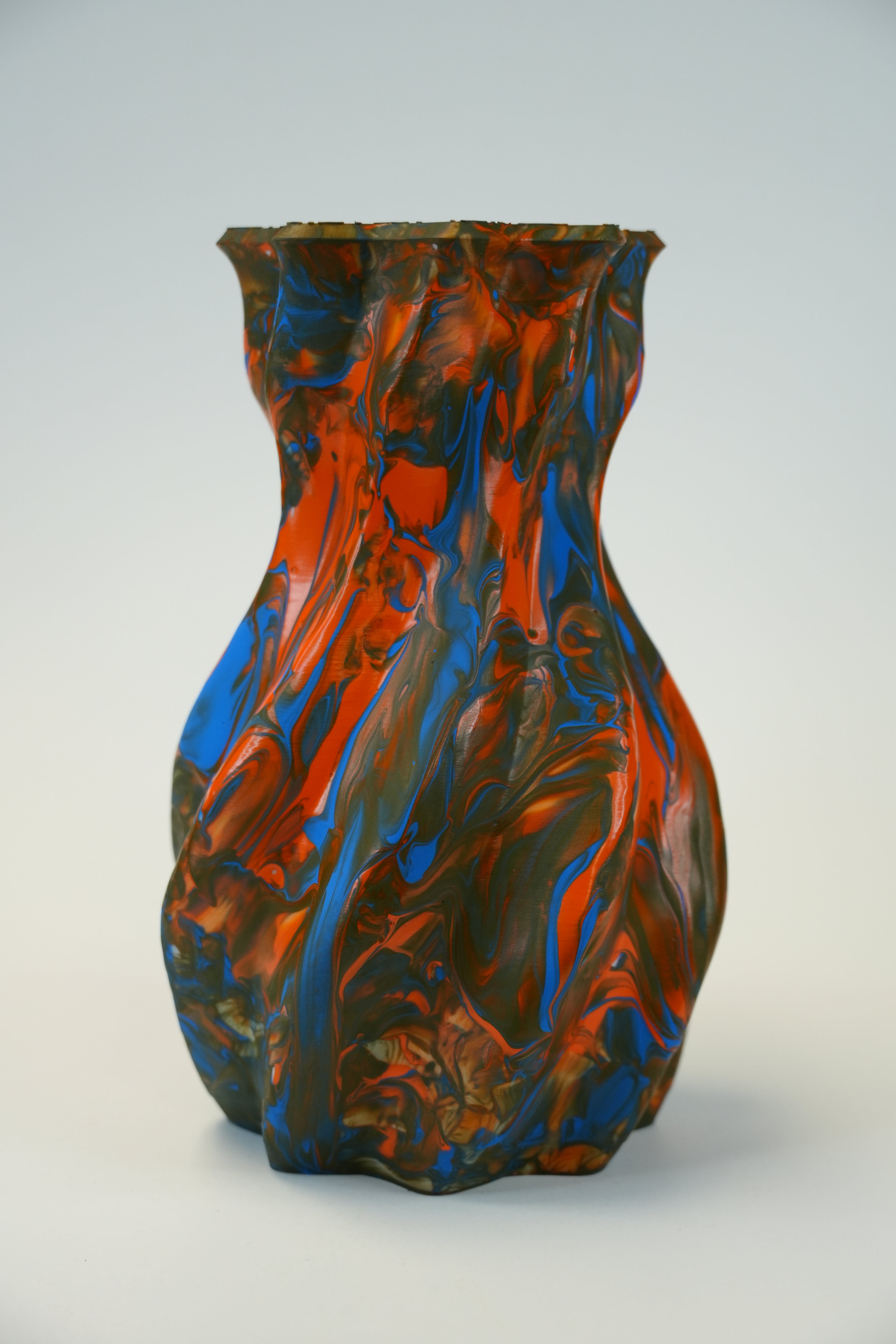A sculptural vase with a flared rim and organic marbled swirl pattern in vibrant blue, orange, and dark brown, set against a neutral light gray background.