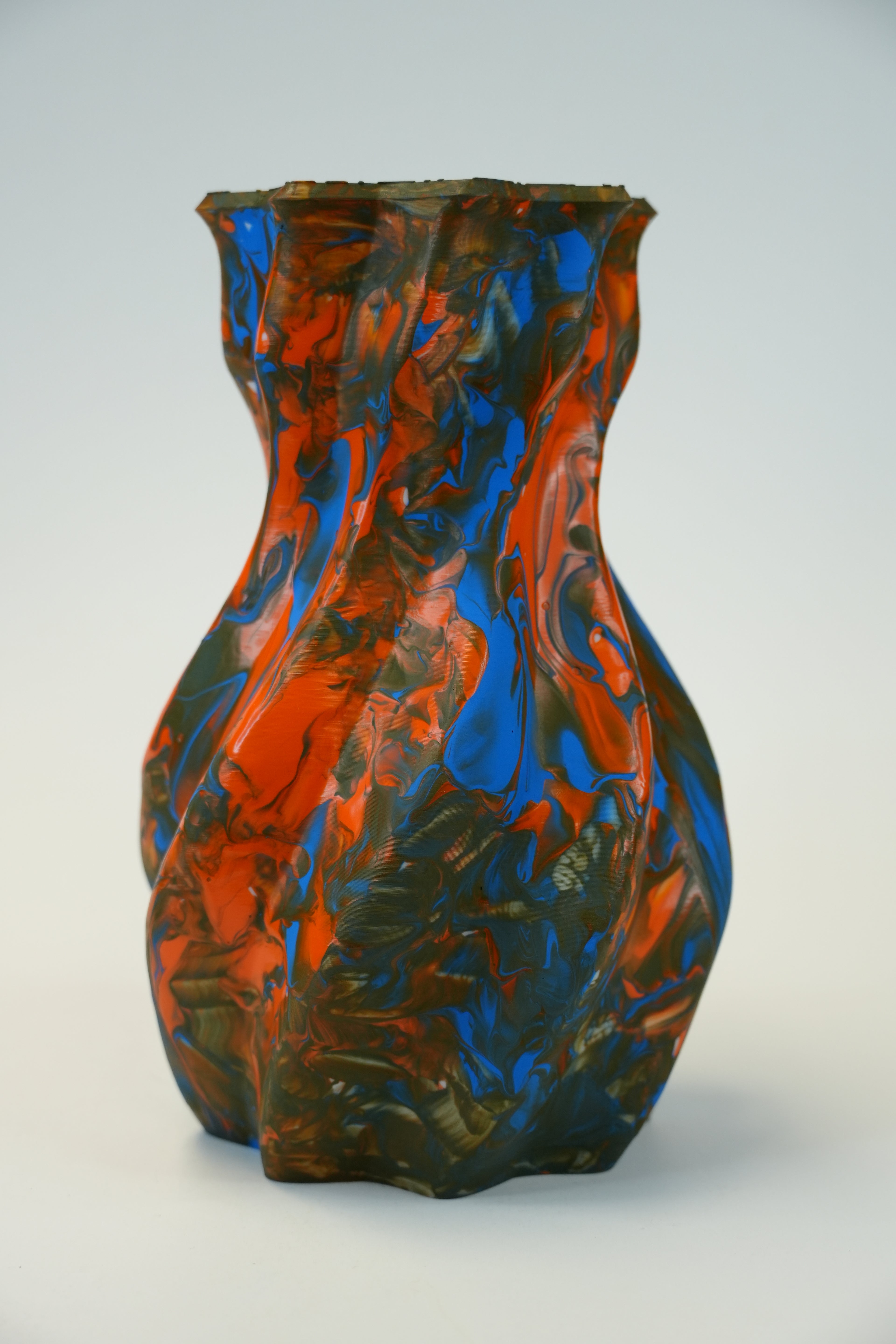 A sculptural, fluted glass vase with a marbled pattern of vibrant orange, deep blue, and dark green, set against a neutral light gray background.