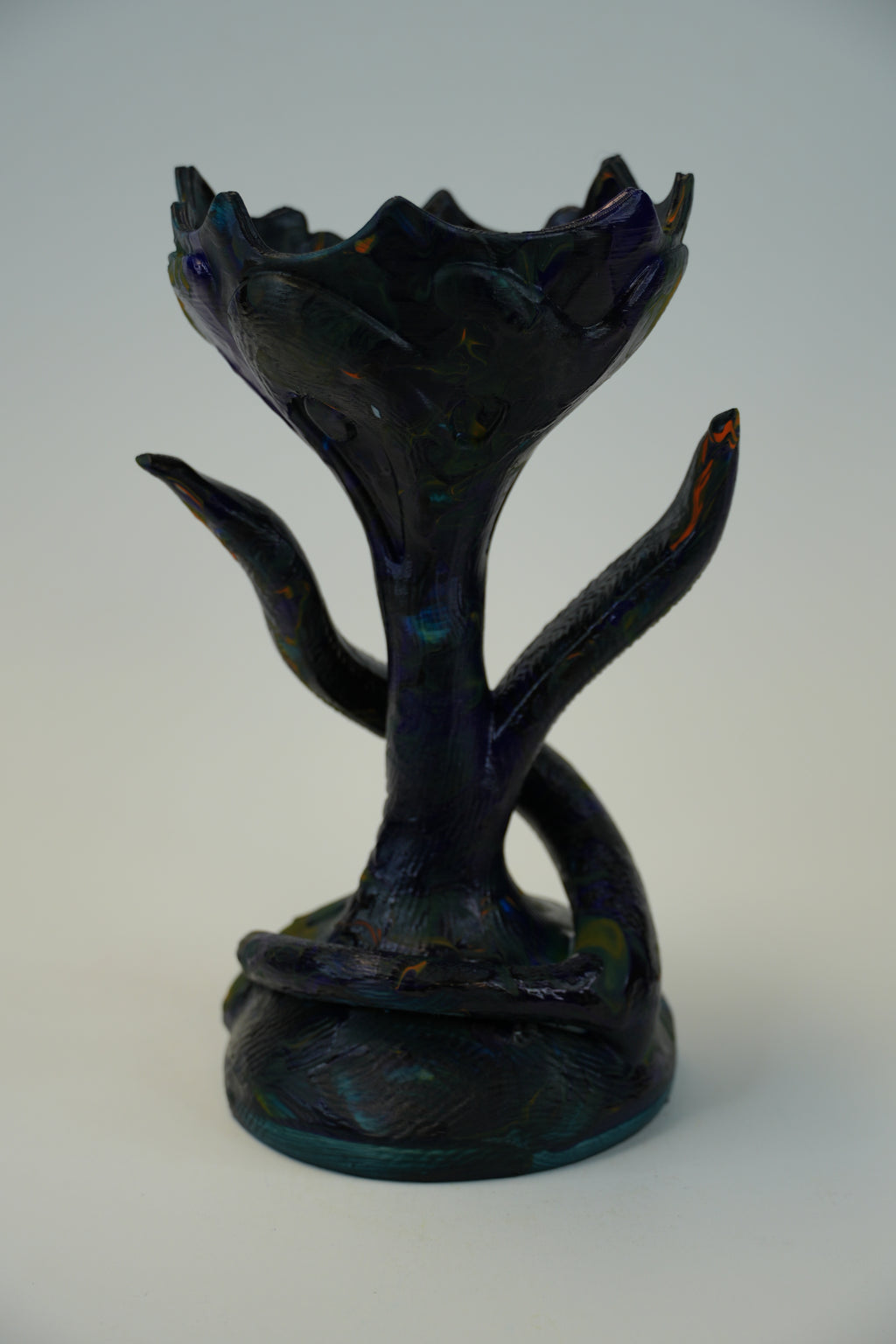A dark iridescent glass sculpture resembling a stylized tree or floral form with branching arms, photographed against a plain light gray studio background.