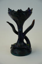 A dark iridescent glass sculpture resembling a stylized tree or floral form with branching arms, photographed against a plain light gray studio background.