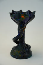 A sculptural glass vase with a dark iridescent body, featuring a wide, wavy rim and a twisted stem that curves around the base, set against a plain light gray background.
