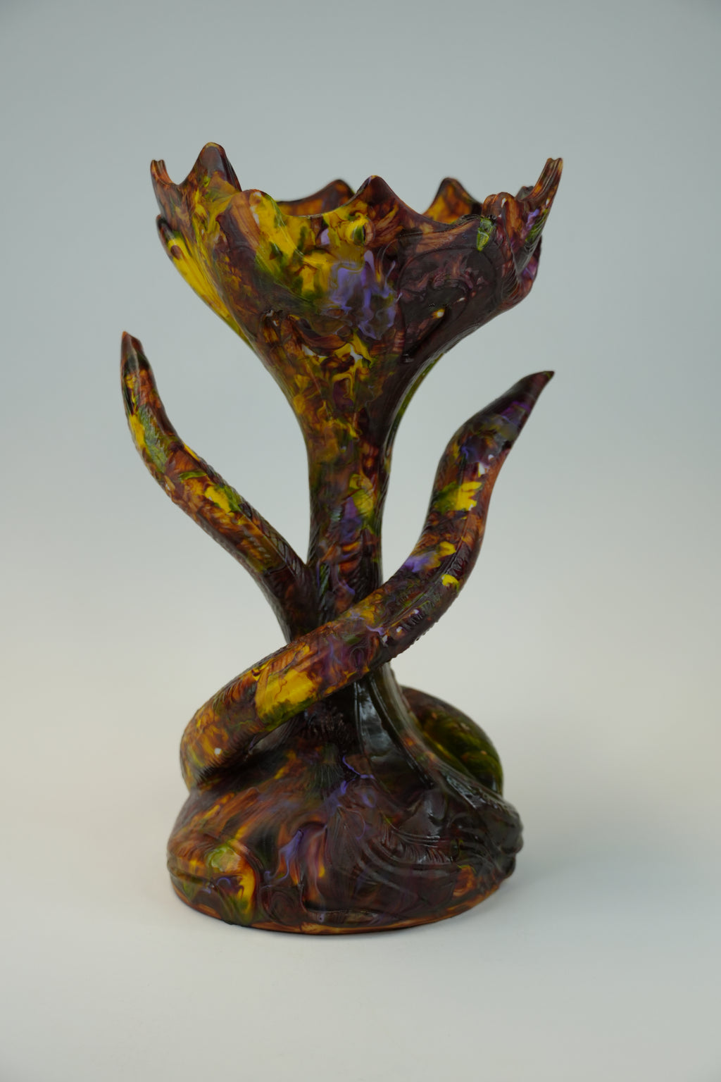A sculptural piece with a stylized tree trunk and flower-bud form, featuring marbled glaze in deep purple, brown, yellow, and green on a light gray studio background.
