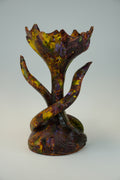 A sculptural piece with a stylized tree trunk and flower-bud form, featuring marbled glaze in deep purple, brown, yellow, and green on a light gray studio background.