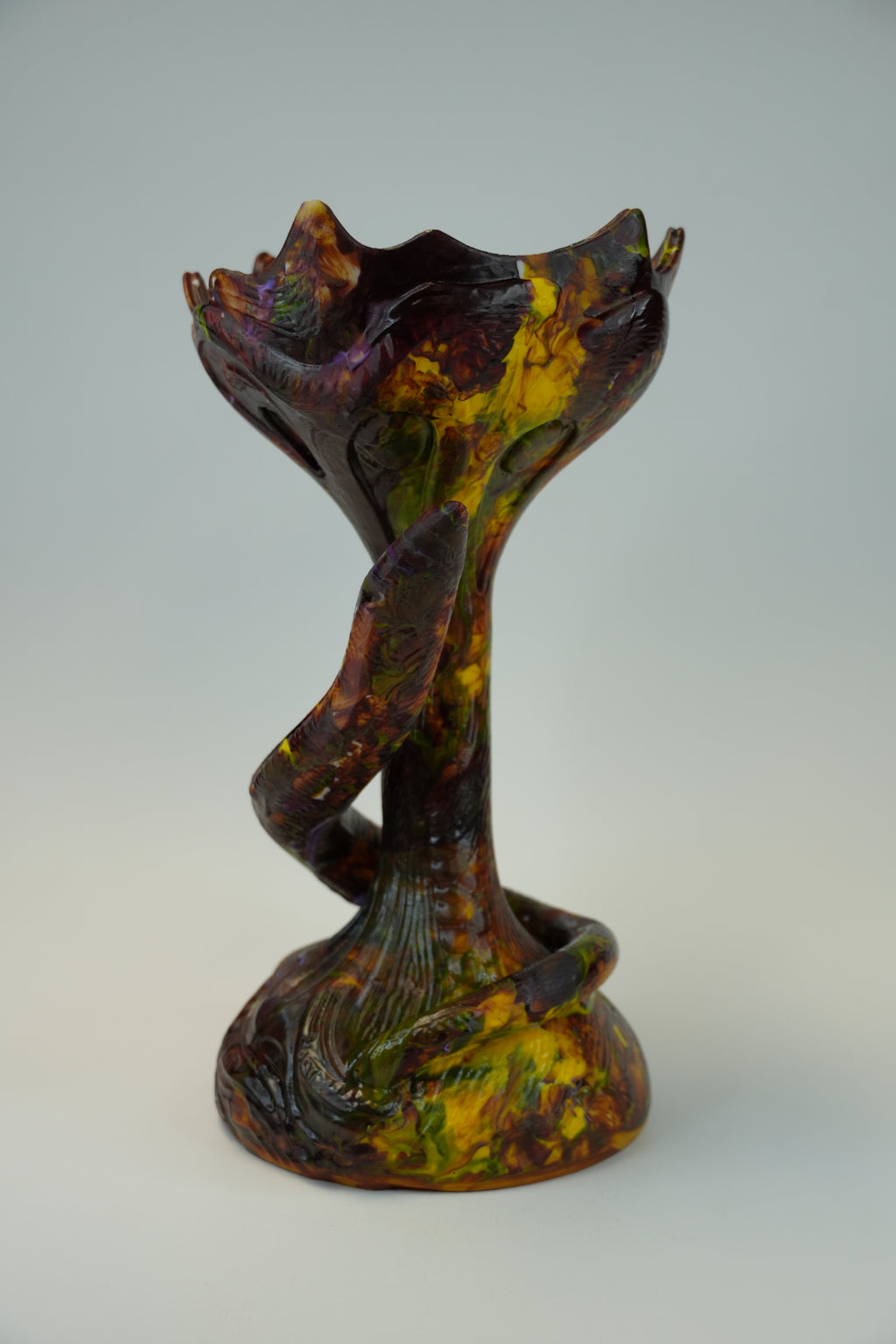An abstract sculptural wall art piece with organic, flowing forms in dark brown, black, yellow, and green tones, mounted against a plain light gray wall.