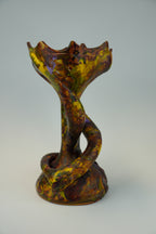An abstract sculptural piece with a twisted, snake-like form and a wide, ruffled rim, featuring a marbled glaze of yellow, green, brown, and purple on a light gray background.
