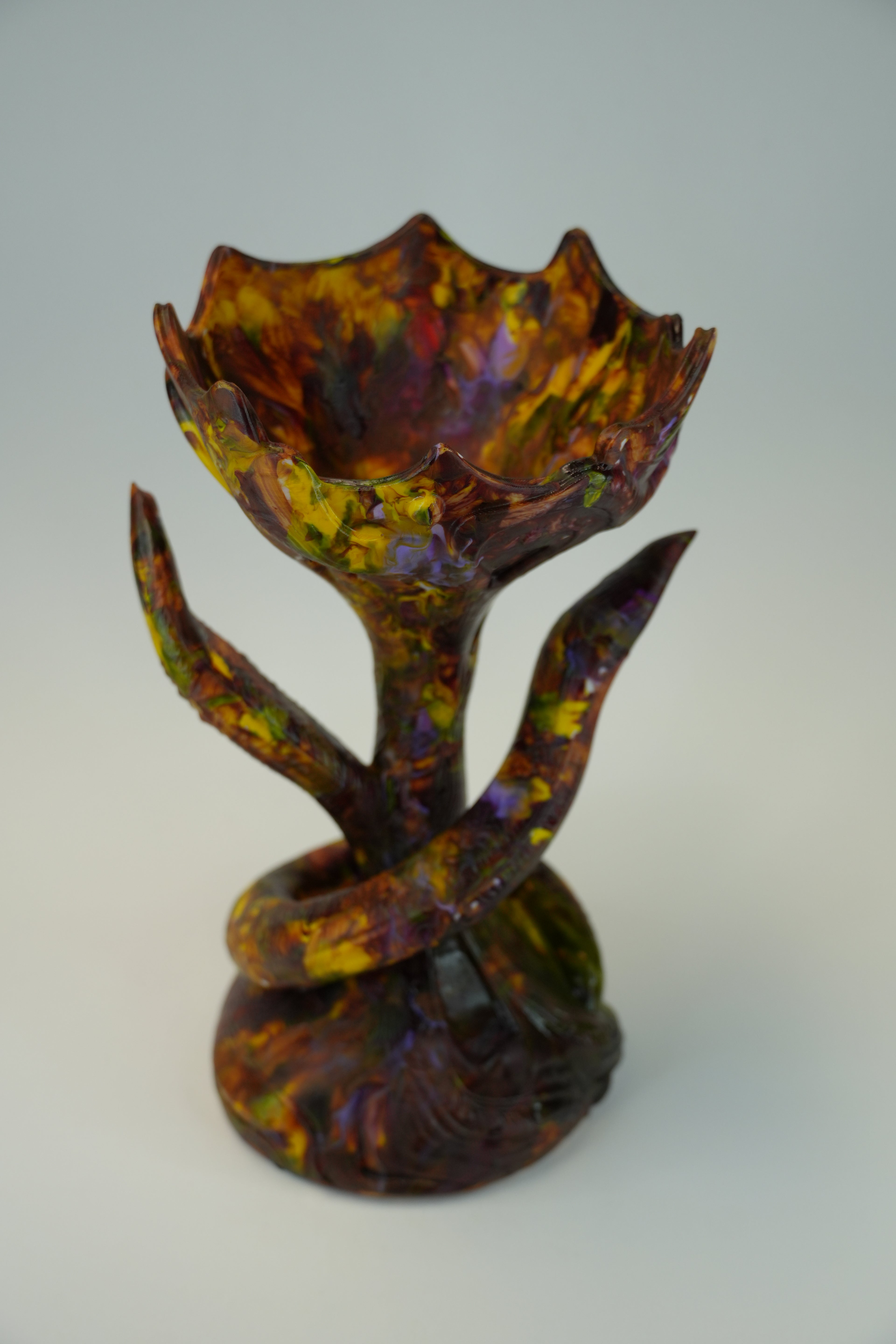 A sculptural piece with a lotus-like open top and twisted stem base, decorated in marbled earthy tones of brown, purple, yellow, and orange on a light gray background.