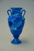 A blue glass urn vase with two loop handles, a fluted neck, and a pedestal base, photographed against a plain light gray studio background.