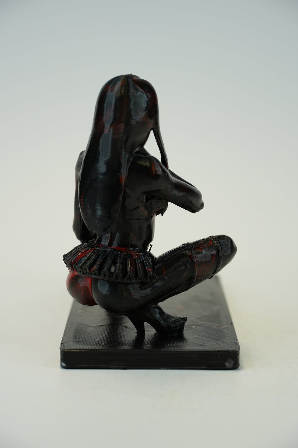 A small black figurative sculpture of a seated woman with long hair, shown from the back on a plain light background.