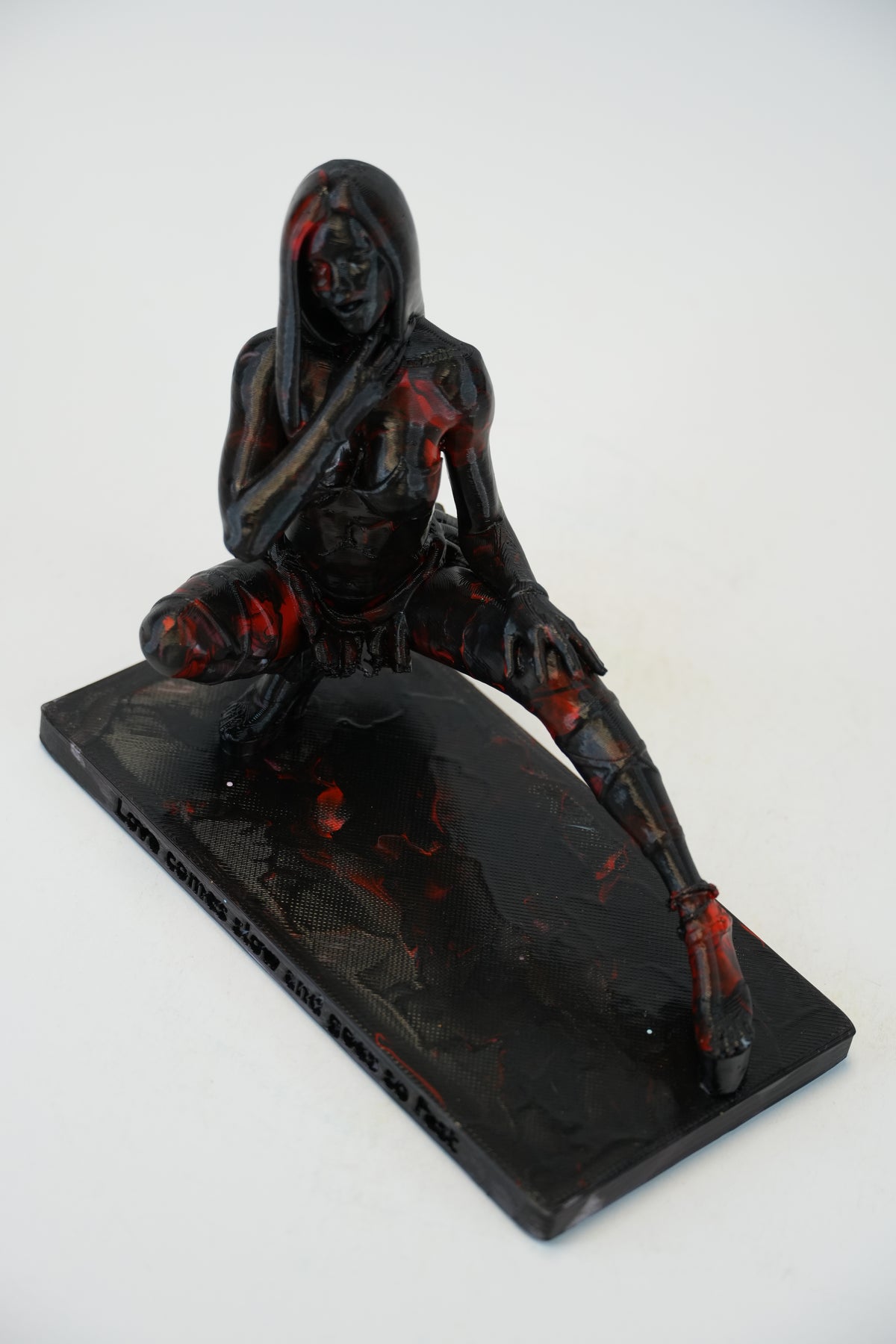 A glossy black resin figurine of a hooded female warrior kneeling on a rectangular base, featuring red and orange marbled inclusions, photographed on a white background.